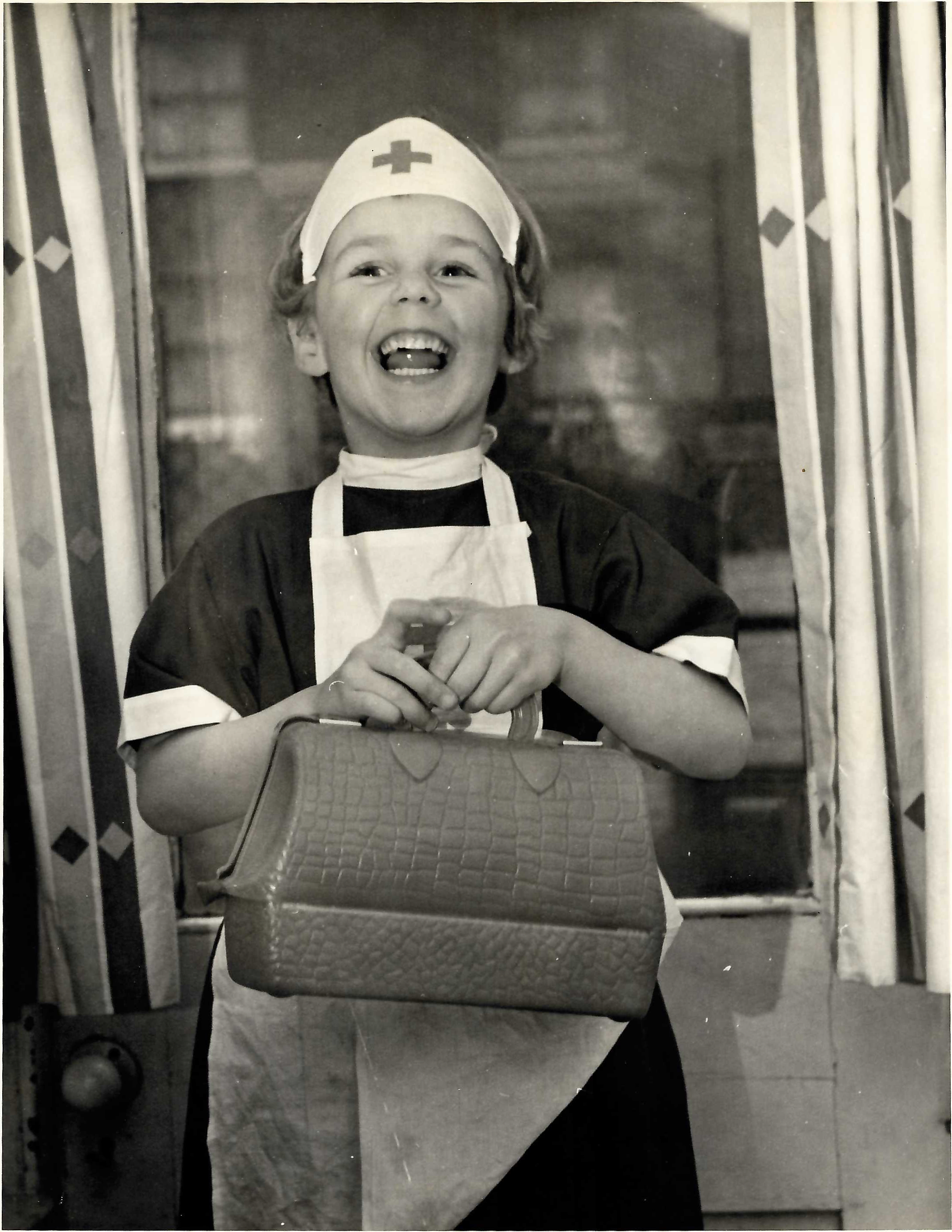 Black and white photo of a young child wearing a nurse's outfit and carrying a toy doctor's bag
