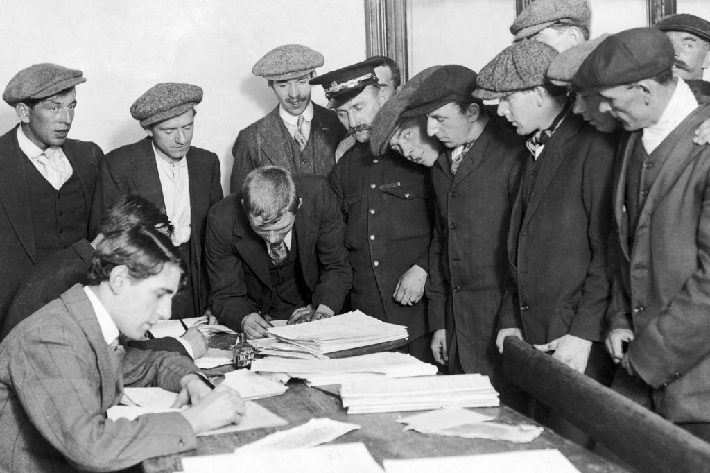 Black and white photo of a group of about 13 young men, mostly in flat caps, queuing to enlist in the British Army in 1914. One man sits at the desk filling out a form and another man leans over the desk signing a form.