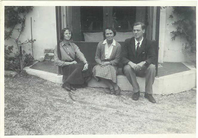 A black and white photograph of a young woman, a middle aged womand and a young man sitting on a step in front of a pair of french windows
