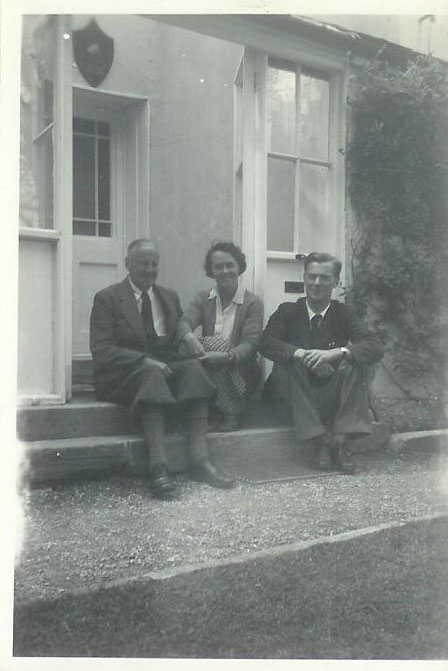 A black and white photograph of an elderly man wearing plus-fours, a middle aged woman and a young man with glasses sitting on a step in front of a conservatory