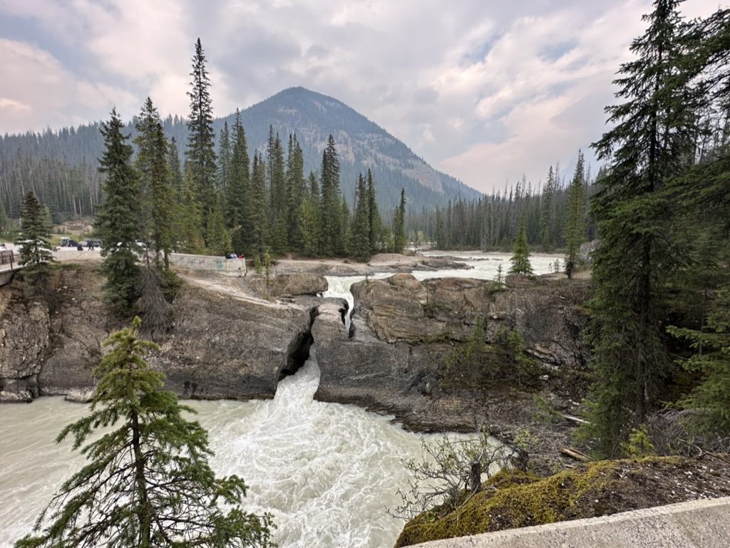 photo of water rushing beneath Natural Bridge, British Columbia, surrounded by pine forests with a mountain in the background