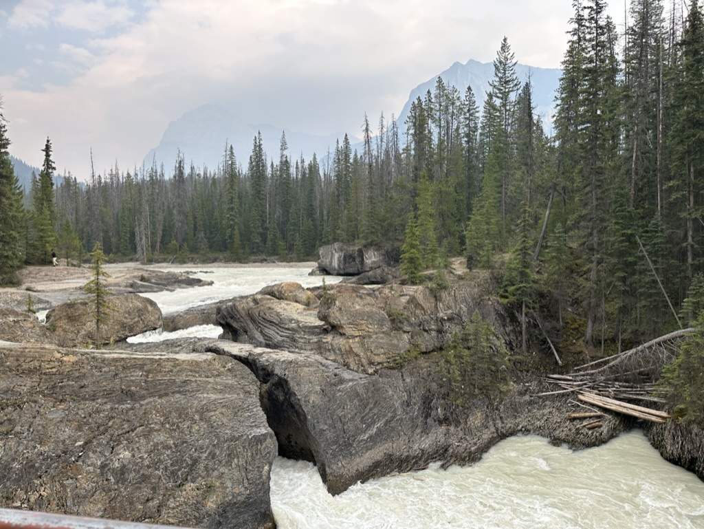 photo of water rushing beneath Natural Bridge, British Columbia, surrounded by pine forests with a mountain in the background