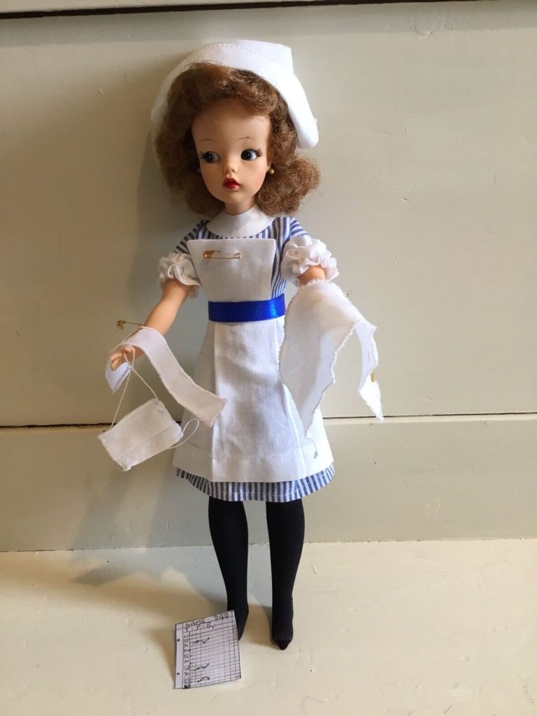 A 'Sindy' doll with dark hair and wearing a nurse's uniform from the 1960s, she is holding bandages and a surgical mask, with an observation chart at her feet.