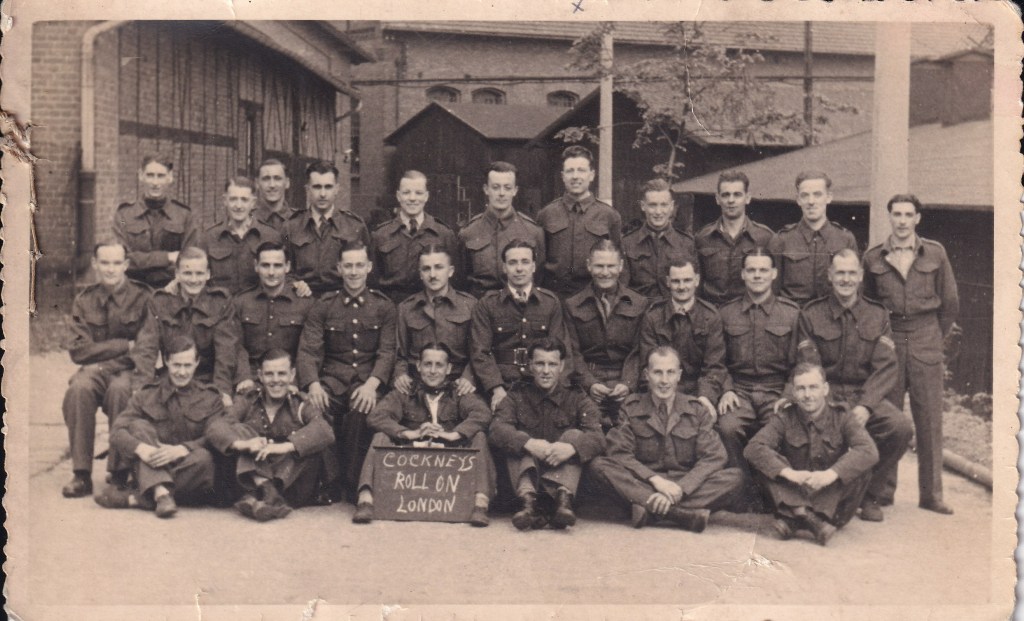 Group photo of WW2 prisoners of war at E72 workcamp attached to Stalag VIIIB POW camp near Lamsdorf. There is a chalkboard caption "Cockneys Roll on London"
