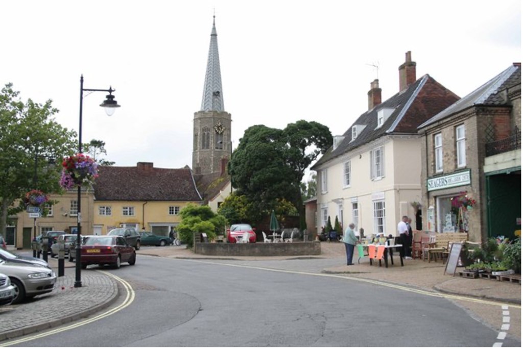 Colour photograph of Wickham Market (c 2015) central square with colourful hanging baskets on the lamposts. All Saints Church with its tall spire is in the background. There are several cars parked around the edge, and a shop with a table outside.