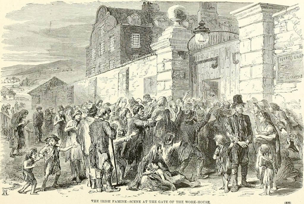 A black-and-white 19th-century illustration titled The Irish Famine—Scene at the Gate of the Work-house. It shows a large, desperate crowd of men, women, and children gathered outside the entrance to a workhouse. Many appear gaunt and in distress, with some reaching out or seated on the ground. The building behind has a heavy stone façade and a sign reading “Work-House.” The image captures the suffering and overcrowding during the Great Irish Famine.