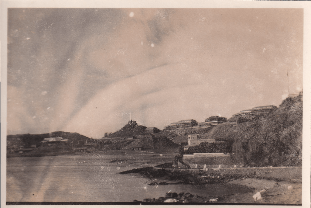 Black and white photograph of Aden, Yemen, showing a coastal landscape with buildings nestled on the hillsides and a cross atop a prominent peak. The foreground features the shoreline and water. c.1945