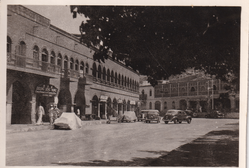 Black and white photograph showing an urban scene in bygone Aden, Yemen, featuring several buildings with arched windows and balconies. A sign reading "DOONG SENG HANG" is visible on one of the buildings. Several vintage cars are parked along the street, and a few people are seen standing or walking nearby.