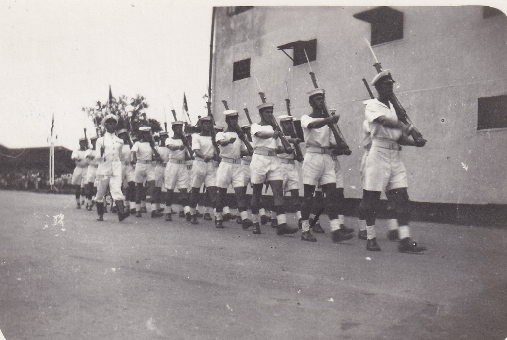 Black and white image of a Royal Naval Guard of Honour in tropical uniform marching in front of a building, the men carry rifles on their shoulders and the officer carries a sword c. 1945