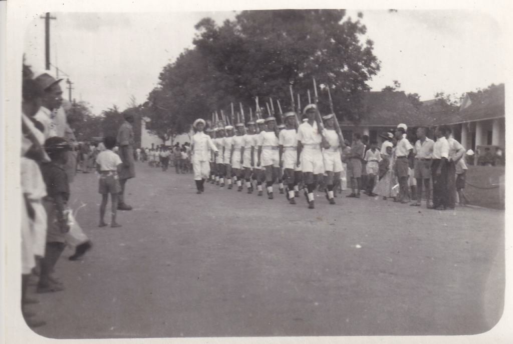 Black and white image of a Royal Naval Guard of Honour in tropical uniform marching. The men carry rifles on their shoulders and the officer carries a sword. The road is lined by standing spectators on either side and there is a low colonial style building in the background c. 1945