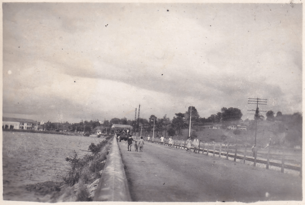 Black and white photograph of the Johor-Singapore Causeway, a 1.056-kilometer structure that serves as a road and rail link between Johor Bahru, Malaysia, and Woodlands, Singapore. There are some pedestrians, one pushing a bicycle. c.1945