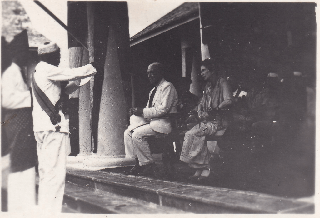 Black and white photograph of a group of people on the steps of a colonial building. A European man wearing a light suit and woman wearing a long dress is beside him. To the left, two men in traditional attire are standing. One of them is gesturing with his right hand.