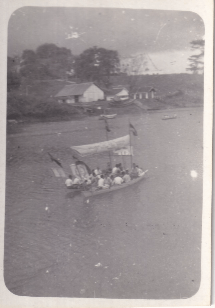 Black and white photograph showing a small boat with flags and a canopy transporting people on a river, likely taken near the Kuching POW camp in Sarawak. Several buildings are visible along the riverbank in the background, suggesting a settlement or camp infrastructure. c.1945