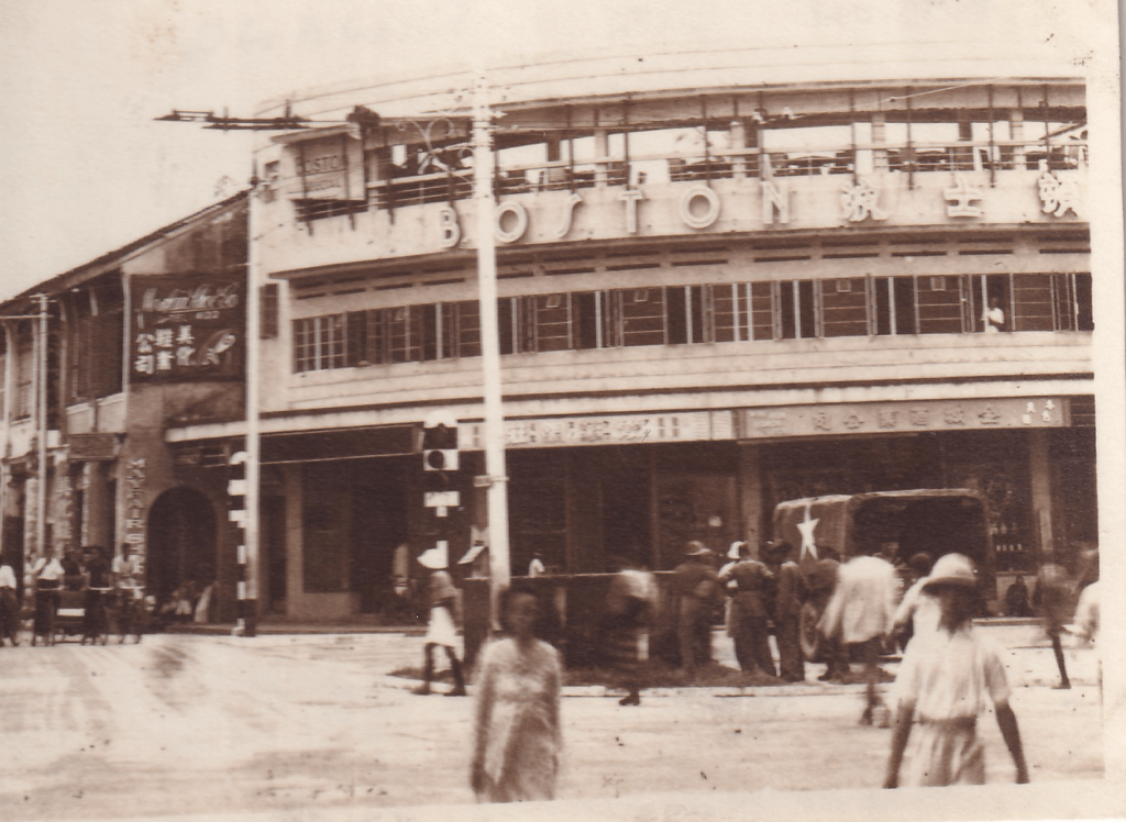 Black and white photograph of the Boston Bar, located at the junction of Penang Road and Prangin Road in George Town, Penang, Malaysia. There are traffic lights, several pedestrians, and an American military vehicle on the street. The building is in Art Deco style featuring the word BOSTON and what appear to be Chinese characters on the facade.