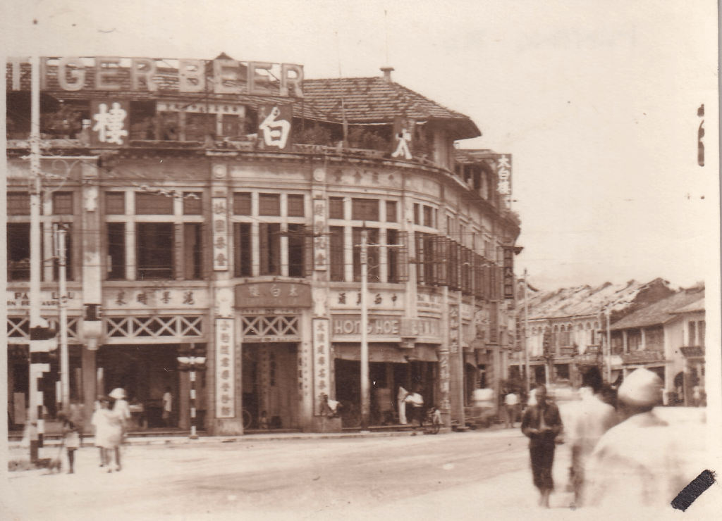 Black and white photograph of the Windsor Building in Penang, Malaysia, The building prominently features the signage "TIGER BEER" and there are several pedestrians on the street. There are traffic lights but no vehicles visible. c.1945