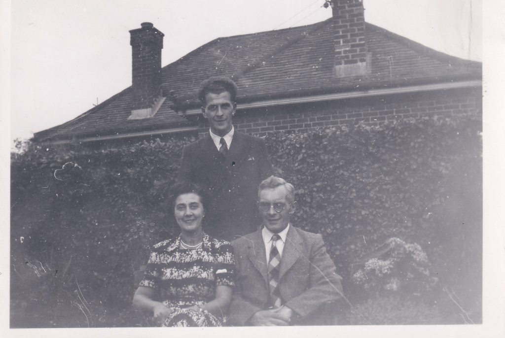 Peter Poulton standing behind his seated parents, Evelyn (‘Sis’) and Stanley Poulton, in a garden in front of a brick house, circa 1940s