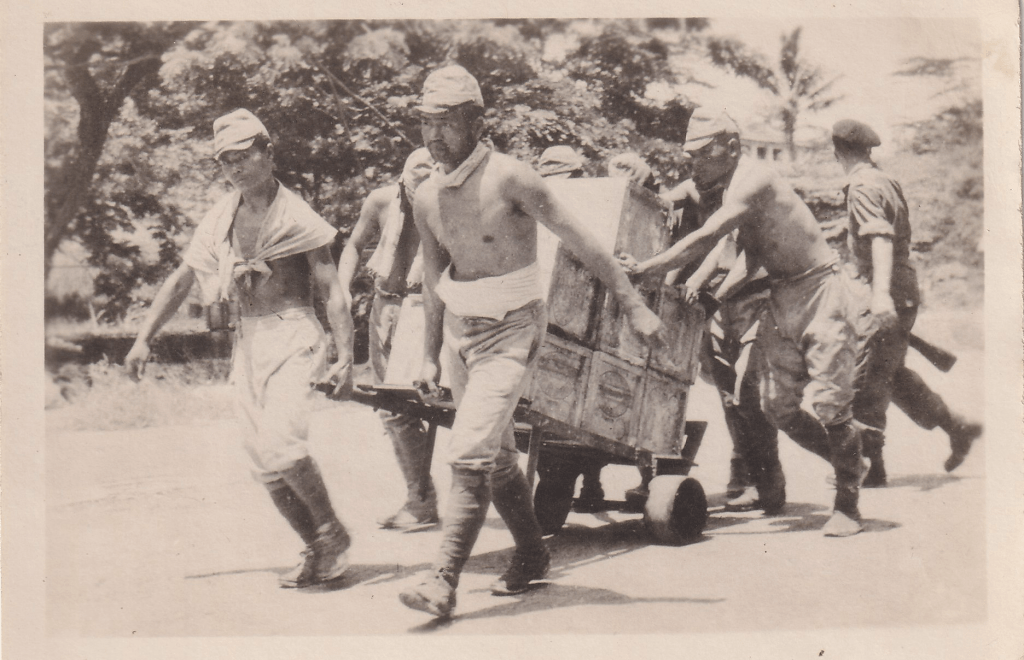 Black and white photograph showing Japanese prisoners of war moving tea chests on a sack truck whilst under armed guard in Singapore