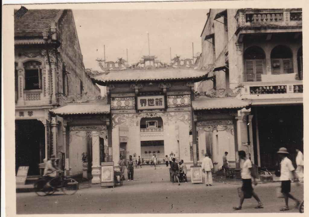 Black and white photograph showing a street scene with pedestrians and someone on a bicycle in front of the gate of the New World Amusement Park, Singapore c.1945