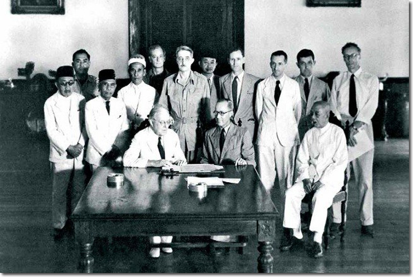 Group of officials, some native but mostly of European appearance, stand behind three men seated.  Two European men sit at a table.  The man on the left in a white suit, is signing some papers whilst the others look on.