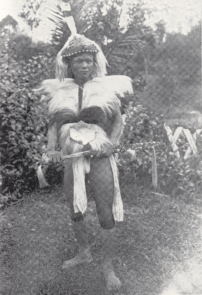 Black and white historic image of a barefoot Mujik hunter from Sarawak dressed in traditional attire including a headdress, clothing decorated with feathers. He appears to be carrying a sword.