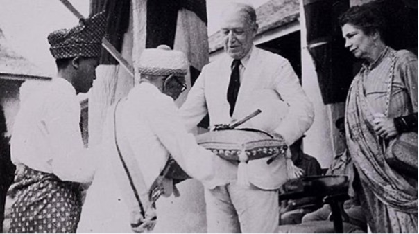 Black and white photograph of a group of people on the steps of a colonial building. A European man wearing a light suit and woman wearing traditional native dress is beside him. To the left, two men in traditional attire are standing. One of them is presenting a ceremonial sword on a cushion. 