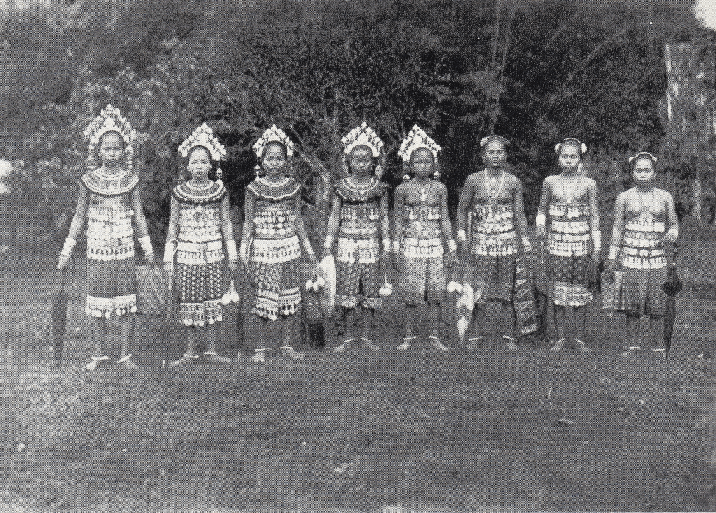 Black and white historic image of eight barefoot Sea Dyak (Iban) girls from Sarawak wearing heavily embellished native dress and holding folded umbrellas. Five are wearing elaborate headdresses and most are bare breasted. The caption states they are carrying smoked heads but none is visible.