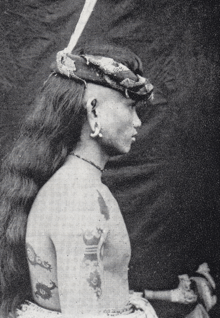 Black and white historic image of a Sea Dyak (Iban) man from Sarawak. His upper body is shown in profile with very long hair, body tattooed, wearing earrings.