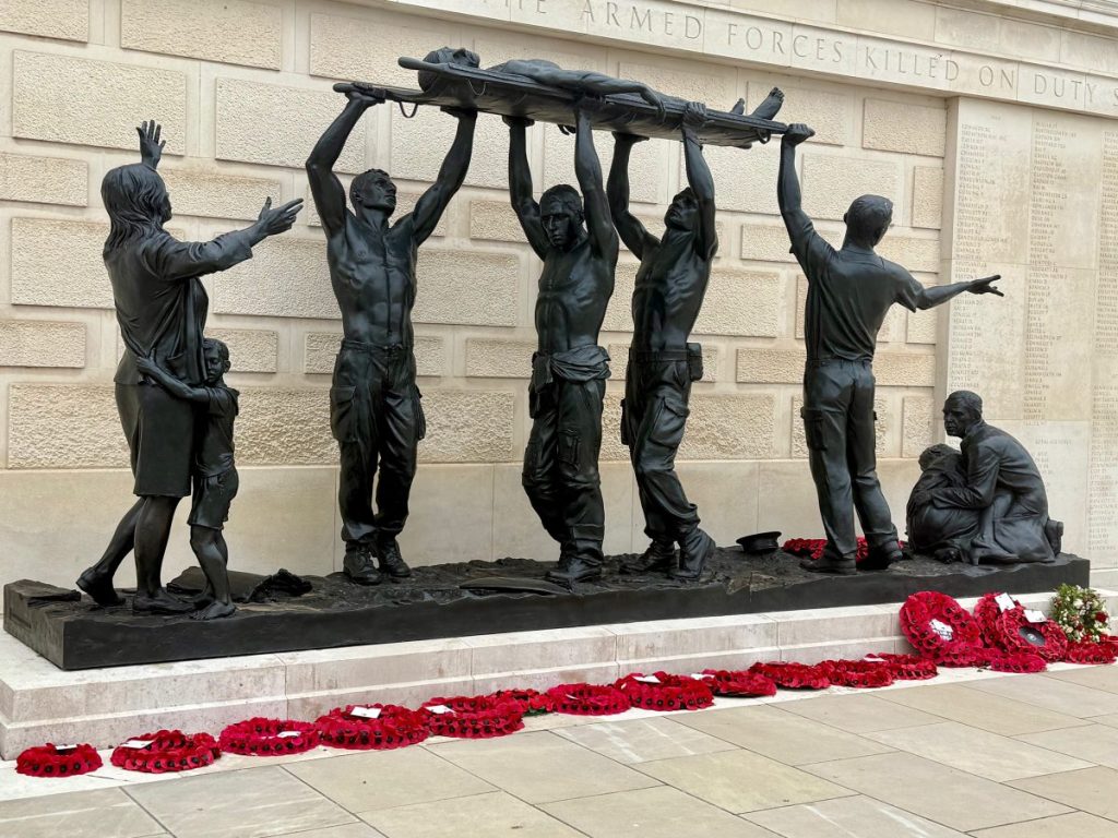 A bronze sculpture depicting several figures, four soldiers carrying a fallen comrade on a stretcherone woman holding a child, and a man comforting another child, with red poppy wreaths laid at their feet. The background features a memorial wall with inscriptions.