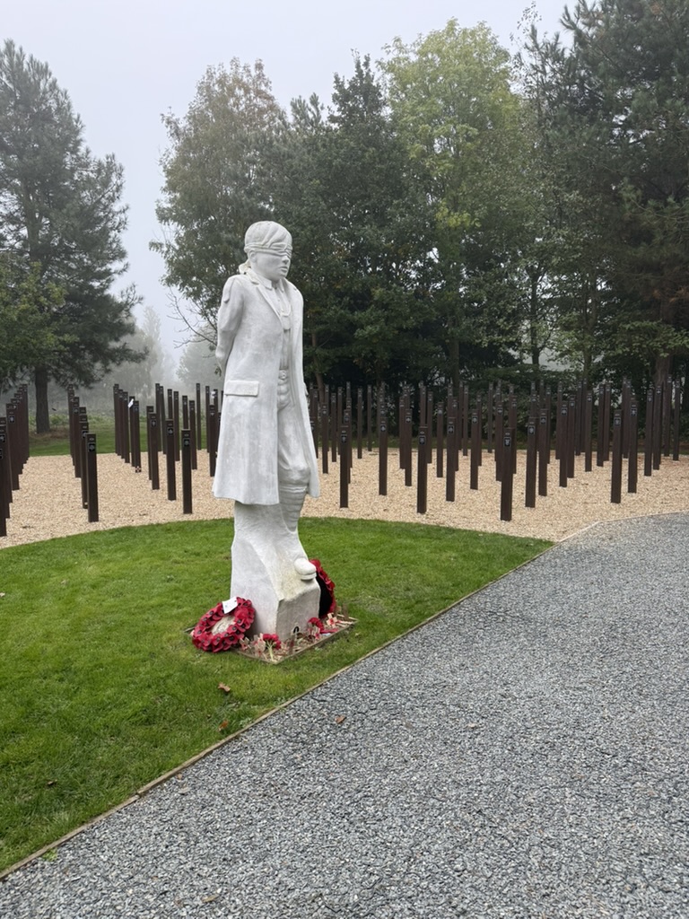 A foggy scene at a memorial site featuring a white statue of a soldier standing on a grassy area, surrounded by rows of wooden stakes, with wreaths placed at the statue's base.