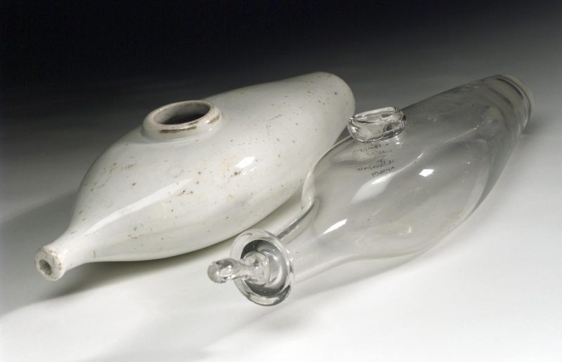 A clear glass infant feeding bottle next to a white ceramic feeding bottle, both shaped with a tapered body and narrow openings.