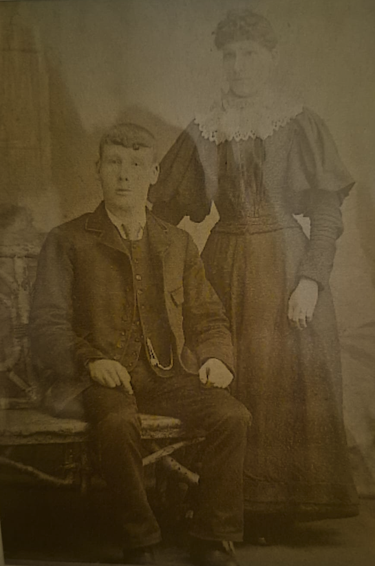 A vintage black and white photograph of a seated man and a woman standing beside him, dressed in late 19th-century clothing. The man is wearing a suit with a waistcoat, while the woman is in a long dress with a high collar.
