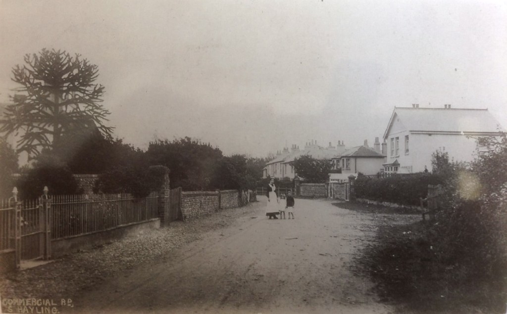 A historic black-and-white photograph of Commercial Road in Hayling, featuring houses and greenery lining the street, with a woman and two children seen walking along the road.