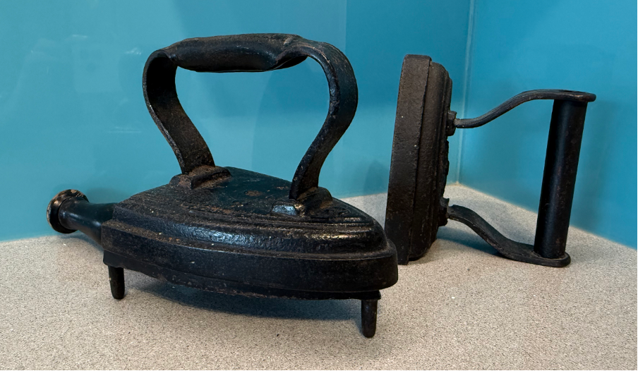 Two antique flat irons on a countertop, one with a curved handle and the other with a flat handle, showcasing vintage laundry tools.