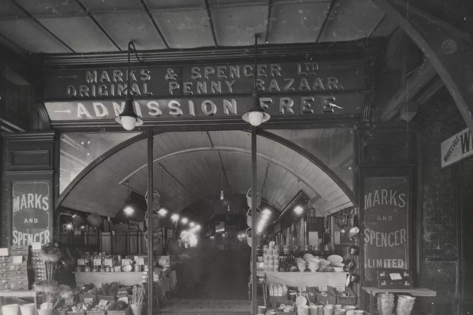 Entrance to the Marks & Spencer Original Penny Bazaar, Brixton, featuring signage that reads 'ADMISSION FREE' and displays of various goods.
