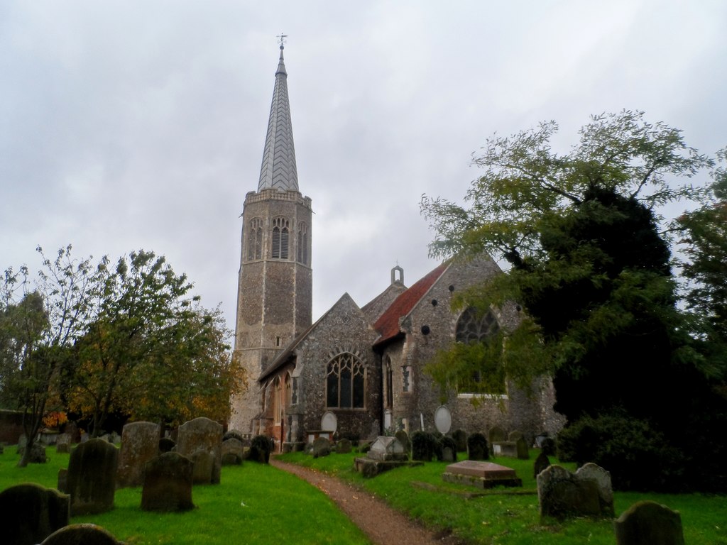 All Saints Church, Wickham Market, a stone church with a tall spire and a graveyard in the foreground, surrounded by trees under a cloudy sky.