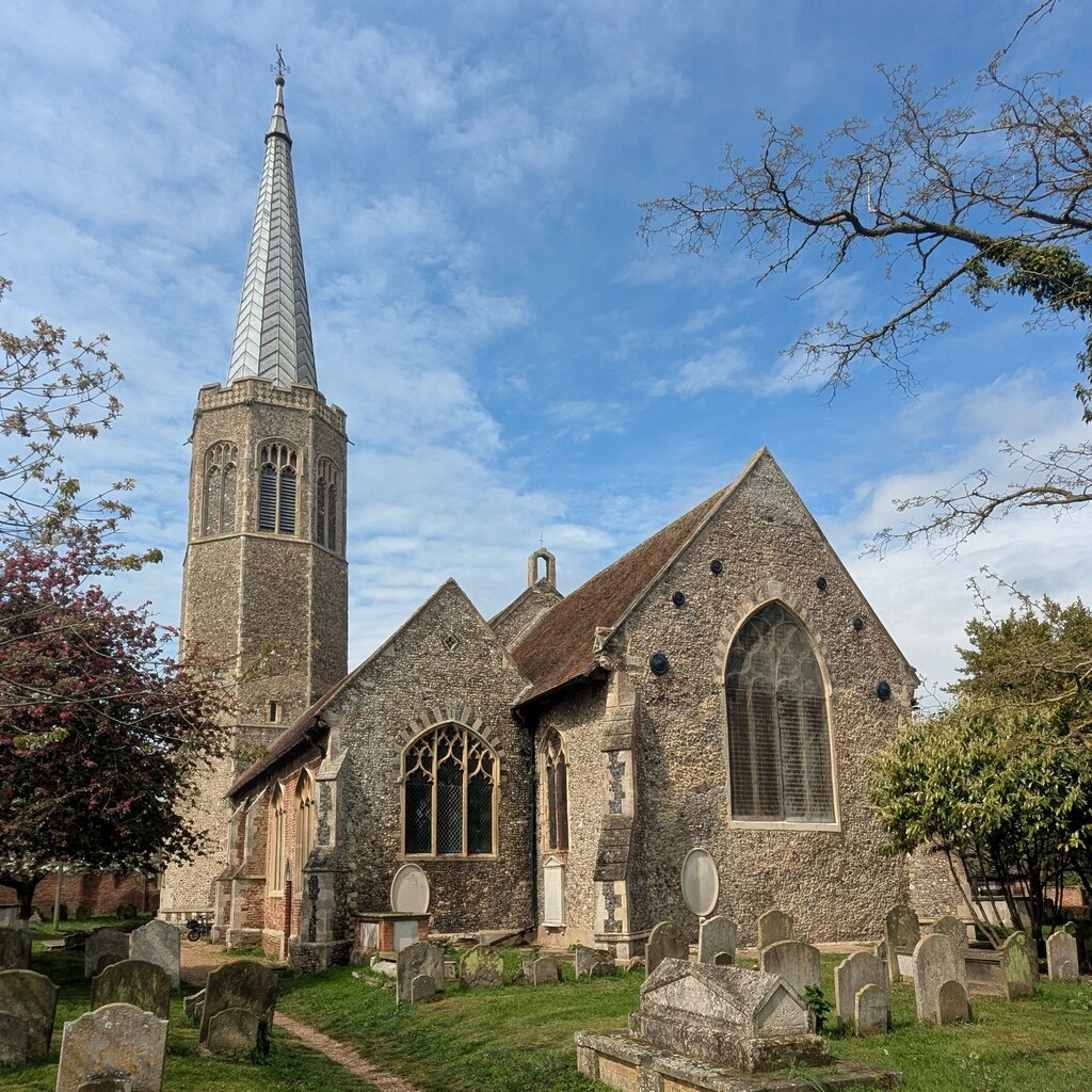 All Saints church, Wickham Market, an historic church building with a tall spire, surrounded by gravestones and lush greenery under a blue sky.