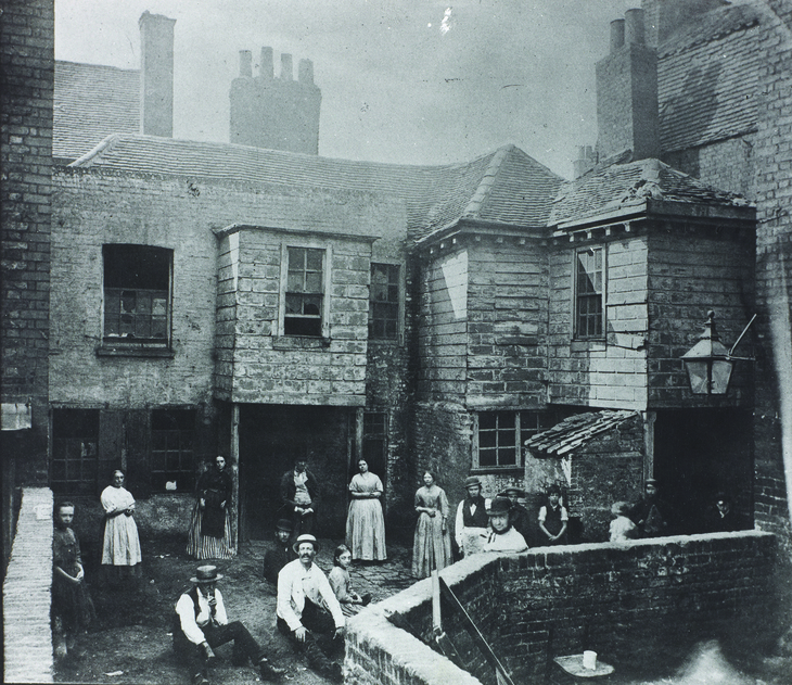 A historic black and white photograph of a group of people standing and sitting outside in a courtyard surrounded by brick buildings, showcasing domestic architecture from the past.