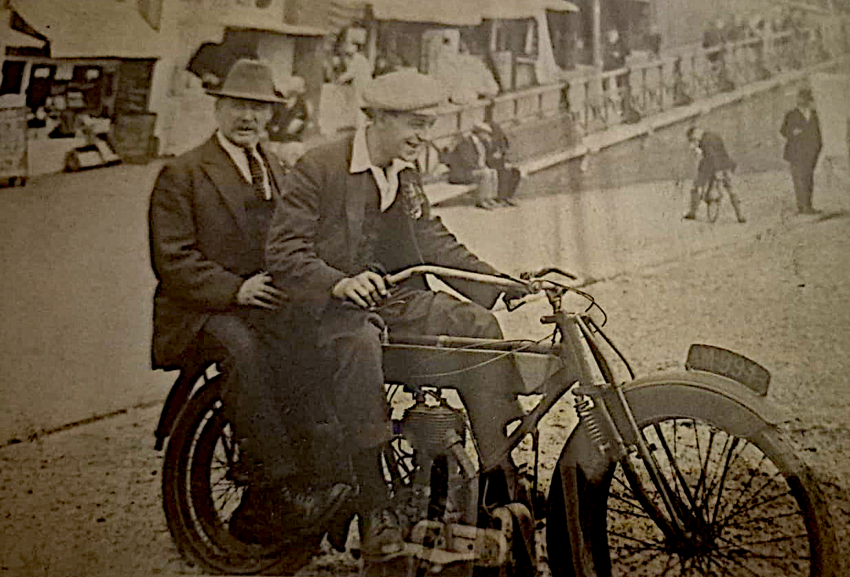 Two men in early 20th century clothing sitting on a vintage motorcycle by a waterfront with people and benches in the background