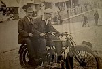 Two men in early 20th century clothing sitting on a vintage motorcycle by a waterfront with people and benches in the background