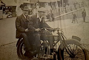 Two men in early 20th century clothing sitting on a vintage motorcycle by a waterfront with people and benches in the background