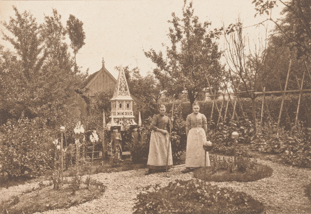 Two women standing on a garden path surrounded by plants and trees, with a decorative wooden structure and garden figures in the background