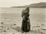 Woman hoeing soil in rocky farmland with baby wrapped on her back near coastline