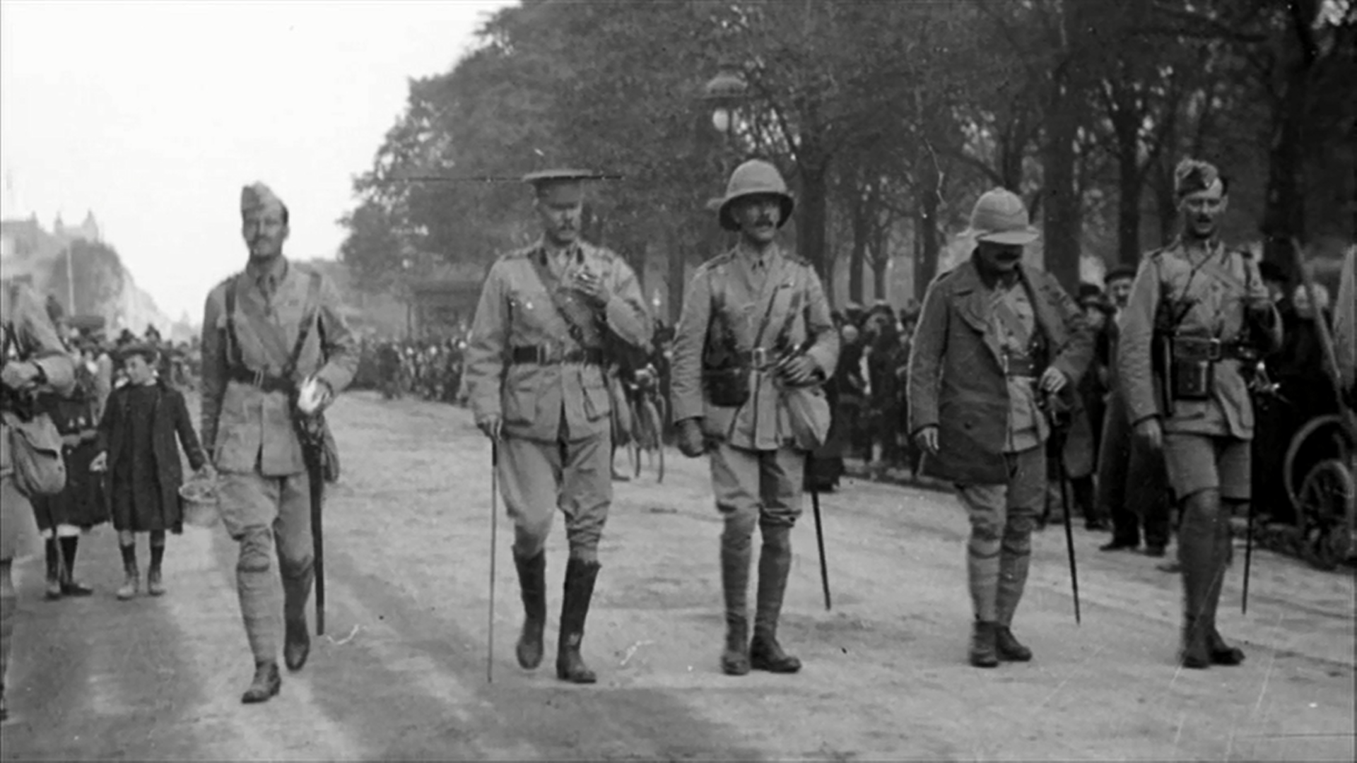 Black-and-white historical photo of uniformed officers walking along a tree-lined road
